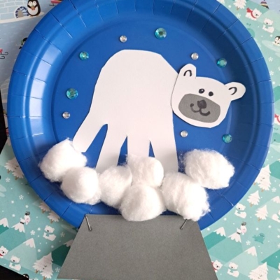 A blue paper plate displays a polar bear made from a white paper handprint, with a drawn face and cotton balls for snow. Decorative sequins and a gray paper iceberg complete the winter craft.