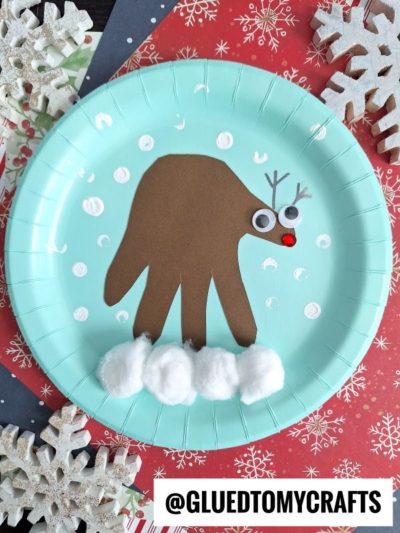 A blue paper plate features a reindeer made from a brown handprint cutout, with googly eyes, a red pom-pom nose, drawn antlers, white paint snow, and cotton balls for snow at the bottom. Snowflakes surround the plate.