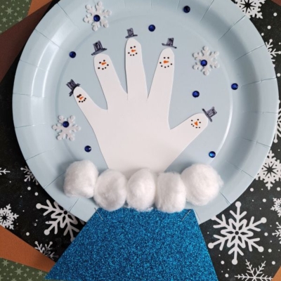 A paper plate craft featuring a white paper cutout of a hand as snowmen, with drawn faces and hats, cotton balls as snow, blue sparkles, and snowflake decorations on a blue background.