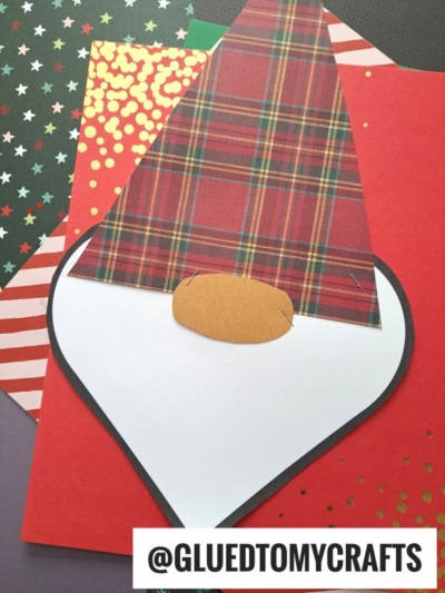 A paper craft gnome with a plaid hat, oval brown nose, and white beard—resembling a heart shaped Santa—is displayed on top of colorful patterned paper. The text @GLUEDTOMYCRAFTS appears at the bottom.