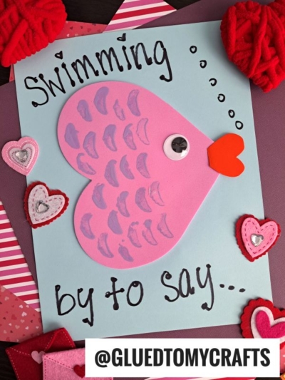 A handmade Valentines Day card features a pink fish made from heart shaped crafts, a googly eye, and a red heart for lips, with Swimming by to say... written on blue paper and heart decorations around.