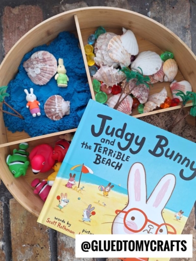A wooden tray with blue sand, seashells, toy bunnies, sea creatures, and palm trees creates the perfect story time sensory bin idea next to the book Judgy Bunny and the Terrible Beach. A @GLUEDTOMYCRAFTS label is displayed.