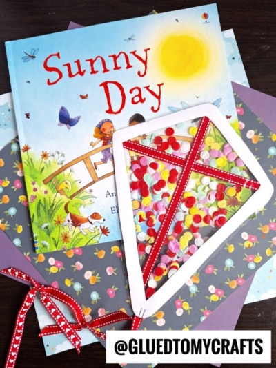 A colorful childrens book titled Sunny Day sits on floral and purple paper, beside a handmade stained glass kite craft filled with confetti and outlined in red with a polka-dot ribbon, with the handle @GLUEDTOMYCRAFTS displayed.