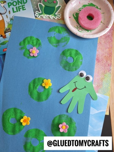 A blue paper displays green stamped lily pads with foam flowers. A green handprint frog with googly eyes and a red smile is on the paper. Craft supplies and a “@GLUEDTOMYCRAFTS” label are nearby.