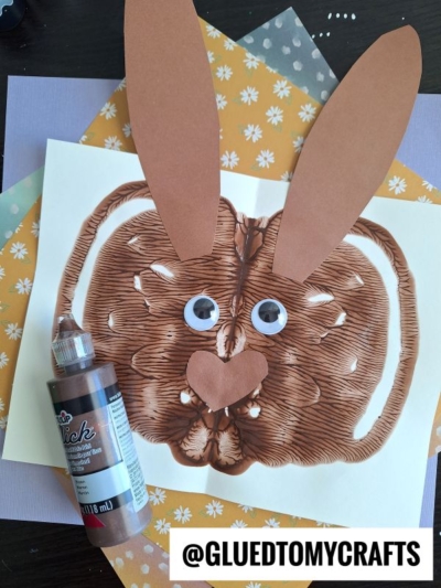 A brown paper bunny paint splat craft with googly eyes and large ears sits on layered pastel papers. A bottle of brown paint and a @GLUEDTOMYCRAFTS label are displayed beside this charming creation.
