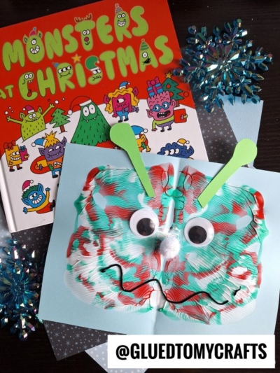 A colorful book titled Monsters at Christmas is shown next to a festive PAINT SPLAT CRAFT featuring googly eyes, a pom-pom nose, and green paper antennae. Snowflake decorations add a wintry touch in the background.