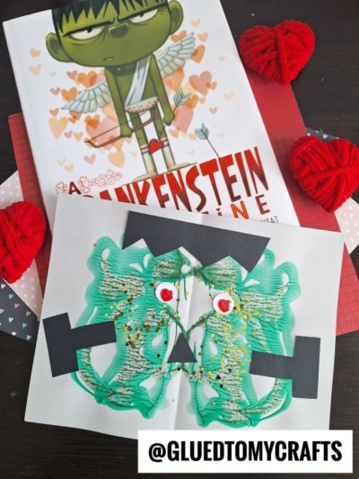 A children’s book titled Frankenstein Valentine sits on a table with red heart decorations and a green, symmetrical PAINT SPLAT CRAFT resembling Frankenstein’s face, made with paint, paper, and googly eyes. @gluedtomycrafts is labeled.