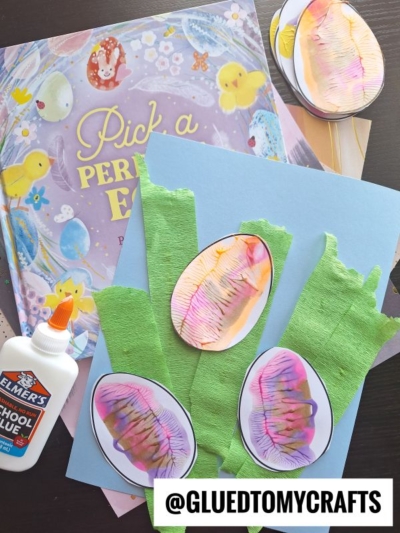 A childrens craft project with watercolor Easter egg cutouts on green tissue paper strips, a bottle of Elmers glue, and an Easter-themed book titled Pick a Perfect Egg. @GLUEDTOMYCRAFTS.