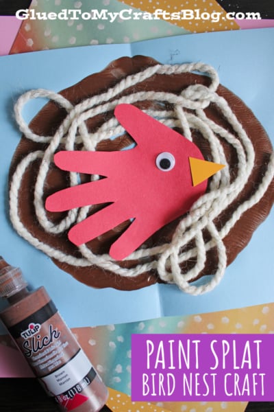 A childrens bird nest craft made with brown paint, white yarn, and a red paper handprint cutout as a bird with a yellow beak. Craft supplies and patterned paper are visible in the background. Text reads: Paint Splat Bird Nest Craft.