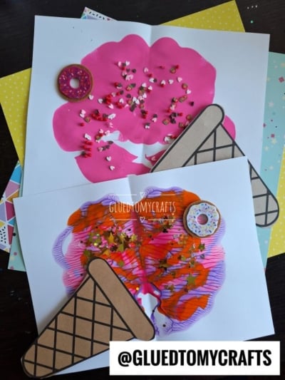 Two pieces of paper display colorful ice cream cone art made with paint and sprinkles. One has a melted ice cream effect with pink paint; the other features pink and orange swirls. Both have paper cone cutouts and are decorated with fake donuts.