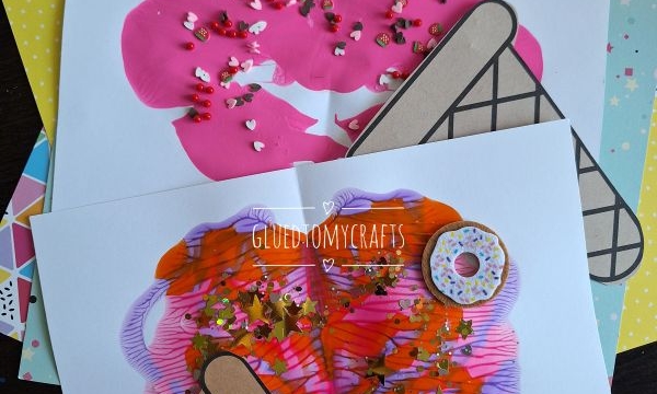 Two pieces of paper display colorful ice cream cone art made with paint and sprinkles. One has a melted ice cream effect with pink paint; the other features pink and orange swirls. Both have paper cone cutouts and are decorated with fake donuts.