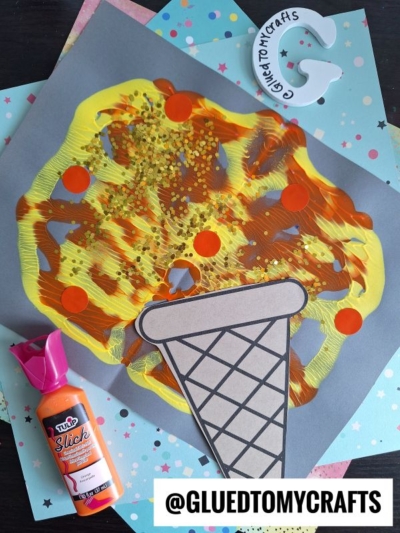 A paper craft of pizza with a cone-shaped crust sits on colorful scrapbook paper. Nearby are orange fabric paint splat, a white letter G with Glued to My Crafts written on it, and a @GLUETOMYCRAFTS label.