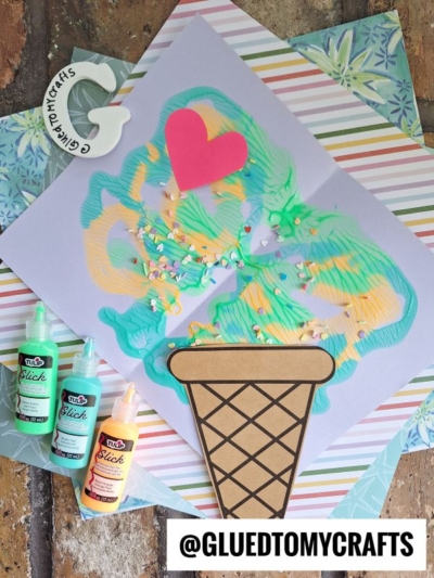 A colorful, paint splat ice cream-themed craft made with swirled paint on paper, decorated with a pink heart, sprinkles, and a paper cone. Tulip paint bottles and craft supplies nearby. @GLUEDTOMYCRAFTS is shown at the bottom.