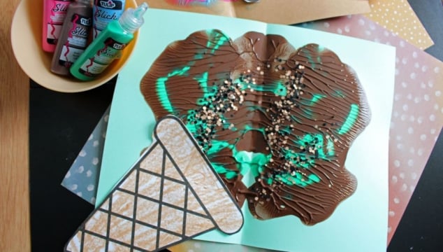 A children’s craft project features an ice cream cone made from paper and painted with swirls of brown, turquoise, and sprinkles. Puffy paint bottles and another similar craft are nearby on a black and polka dot surface.