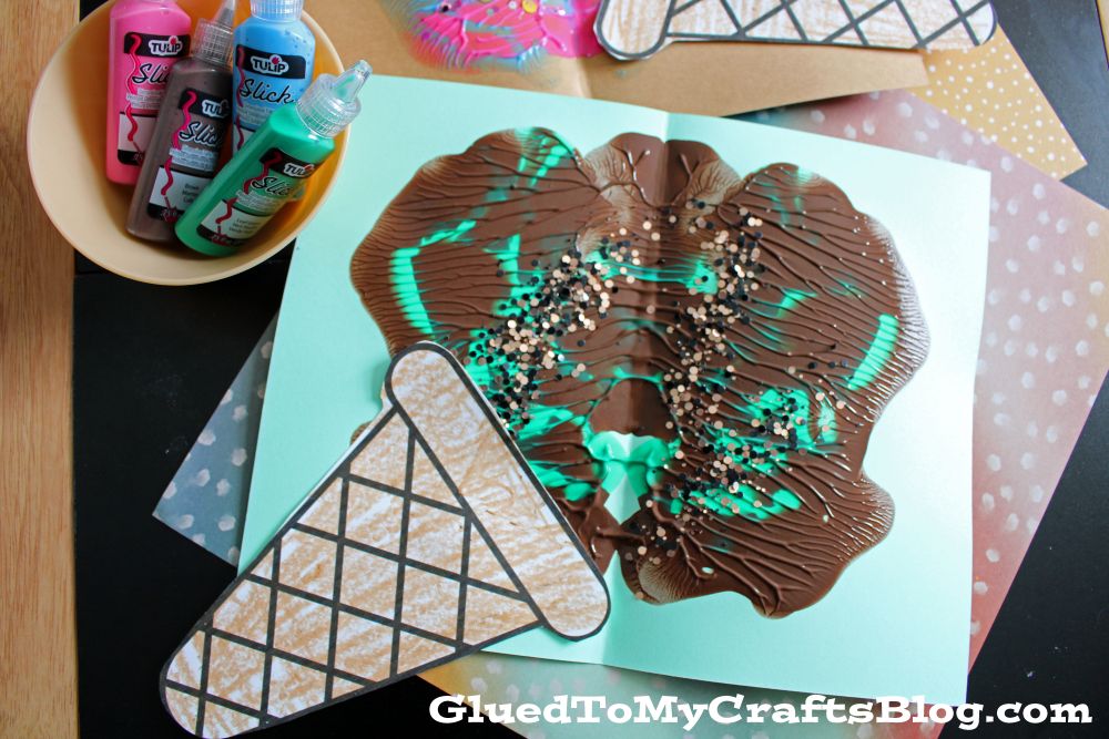 A children’s craft project features an ice cream cone made from paper and painted with swirls of brown, turquoise, and sprinkles. Puffy paint bottles and another similar craft are nearby on a black and polka dot surface.