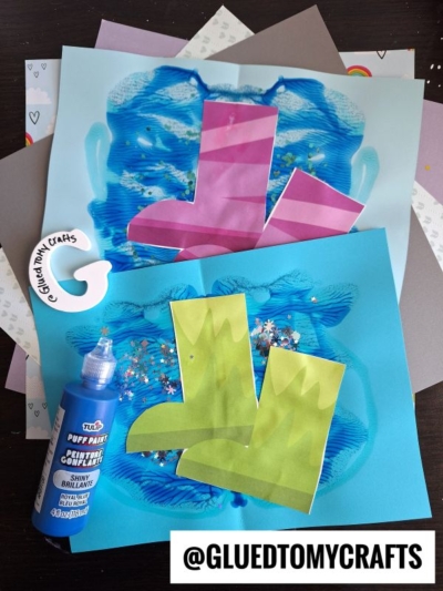 Colorful childrens craft project featuring cut-out paper rain boots on blue, paint-splattered backgrounds, a bottle of blue puff paint, glitter, and a “@GLUEDTOMYCRAFTS” handle in the corner.
