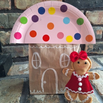 A gingerbread house craft made from a brown paper bag decorated with white chalk, topped with a pink paper plate roof covered in colorful dots. A small gingerbread doll in a red dress stands beside it.