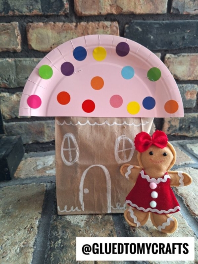A gingerbread house craft made from a brown paper bag decorated with white chalk, topped with a pink paper plate roof covered in colorful dots. A small gingerbread doll in a red dress stands beside it.
