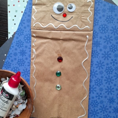 A paper bag gingerbread man is decorated with googly eyes, a red pom-pom nose, white icing outlines, and colorful gem buttons. Nearby are a glue bottle, craft paper, and extra googly eyes. Text: @GLUEDTOMYCRAFTS.