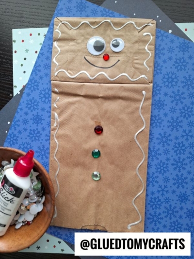 A paper bag gingerbread man is decorated with googly eyes, a red pom-pom nose, white icing outlines, and colorful gem buttons. Nearby are a glue bottle, craft paper, and extra googly eyes. Text: @GLUEDTOMYCRAFTS.