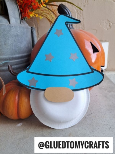 A paper plate craft shaped like a wizard’s face, similar to a paper bowl wizard, features a blue hat with silver stars and a tan nose, displayed in front of pumpkins. Text at the bottom reads @GLUEDTOMYCRAFTS.