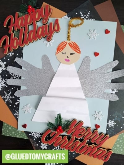 A Christmas craft features a paper angel with handprint wings, surrounded by snowflakes, hearts, and festive Happy Holidays and Merry Christmas signs, with a green @GLUEDTOMYCRAFTS tag at the bottom.