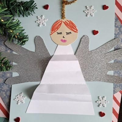 A handmade angel craft features a paper angel with glittery silver wings, a gold pipe cleaner halo, and a white dress. It is surrounded by snowflake stickers, red hearts, and pine branches on a blue background.