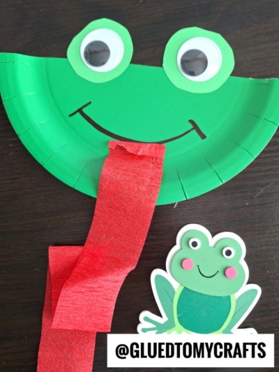 A green paper plate frog craft with large googly eyes, a smiling black mouth, and a long red paper tongue, next to a cartoon frog sticker and @GLUEDTOMYCRAFTS text at the bottom.
