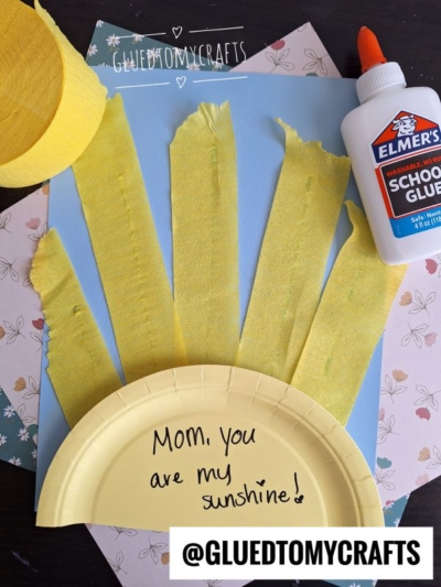 A paper plate with Mom, you are my sunshine! is decorated with yellow crepe paper strips, school glue, and scrapbook paper—one of the sweetest crepe paper craft ideas. The handle @GLUEDTOMYCRAFTS is visible at the bottom.