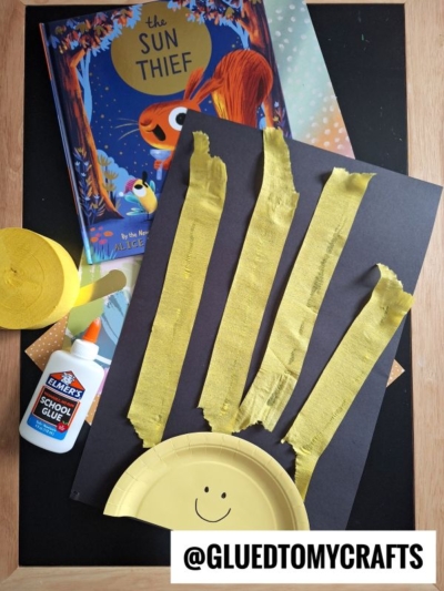 A children’s crepe paper craft idea: black paper with yellow strips and a half yellow paper plate forming a sun, next to The Sun Thief book, Elmer’s glue, yellow crepe paper, and a @GLUEDTOMYCRAFTS label.