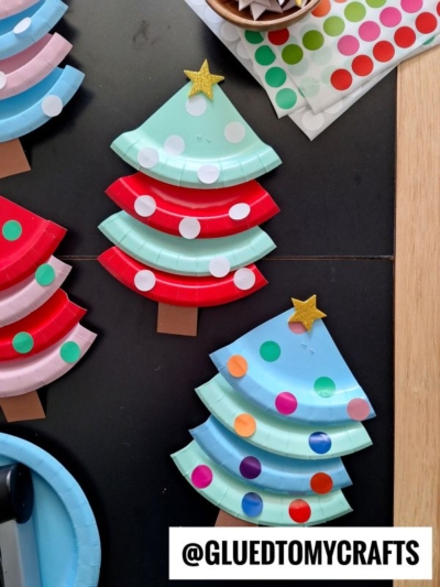 Three Christmas tree crafts made from painted paper plates decorated with colorful dot stickers and topped with gold stars. The trees have brown paper trunks and are displayed on a black surface.