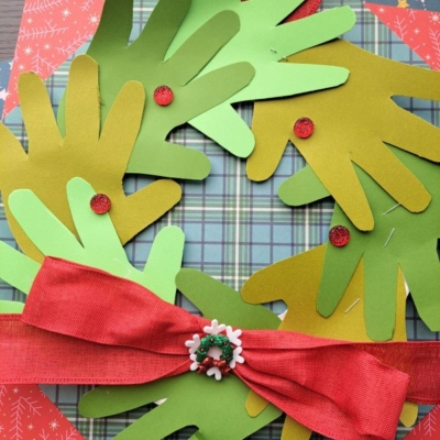A handprint Christmas wreath made from green paper cutouts, decorated with red dots and a red bow, is displayed on festive patterned paper. The handle @GLUEDTOMYCRAFTS appears at the bottom.