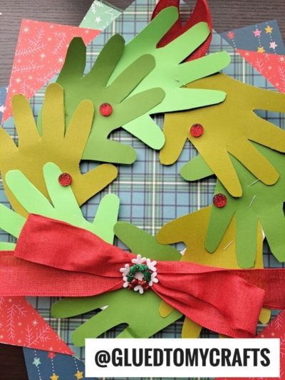 A handprint Christmas wreath made from green paper cutouts, decorated with red dots and a red bow, is displayed on festive patterned paper. The handle @GLUEDTOMYCRAFTS appears at the bottom.