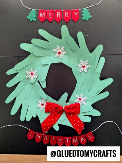 A handmade Christmas wreath made from green paper hand cut-outs, decorated with white snowflakes and a red bow. Above and below are red felt banners reading Merry Christmas with green tree shapes.