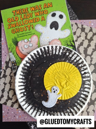 A children’s book titled “There Was an Old Lady Who Swallowed a Ghost!” next to a paper plate craft featuring a yellow moon, a small ghost cutout, and star decorations. The handle @GLUEDTOMYCRAFTS is at the bottom.