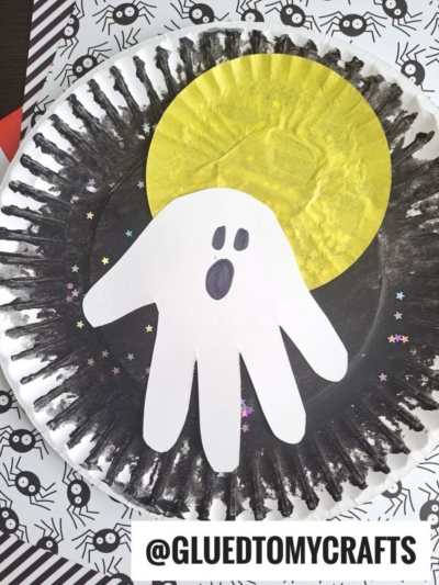 A paper plate painted black with a yellow circle for a moon and a white handprint ghost in the center, decorated with star-shaped confetti. The background has a spider web pattern. Text at bottom reads @gluedtomycrafts.