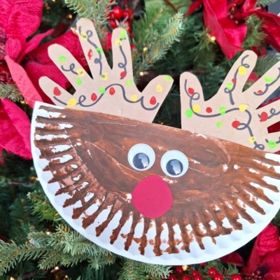 A handmade reindeer ornament with a brown painted paper plate face, googly eyes, a red paper nose, and paper handprint antlers decorated with colorful light designs, hanging on a Christmas tree with red poinsettias.