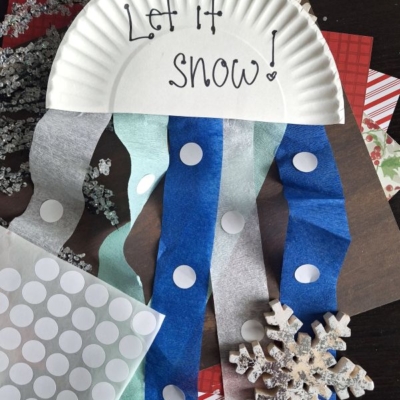 A paper plate with Let it snow! written on it is decorated with blue and white paper streamers and white dot stickers to resemble a snow-themed craft. Snowflake decorations and patterned papers are nearby. @GLUEDTOMYCRAFTS is at the bottom.