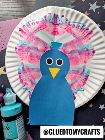 A PAINT SPLAT CRAFTS project featuring a blue paper bird with googly eyes and a yellow beak on a paper plate decorated with pink and blue symmetrical handprint splats, surrounded by art supplies and star-patterned paper.