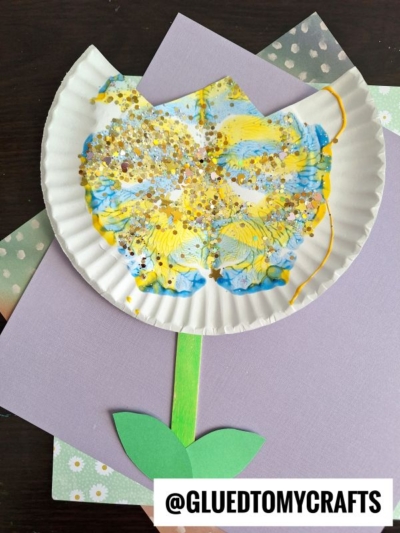 A paper plate painted with blue and yellow, shaped like a flower, decorated with gold glitter. It has a green paper stem and leaf, and is placed on layered pastel-colored paper backgrounds.