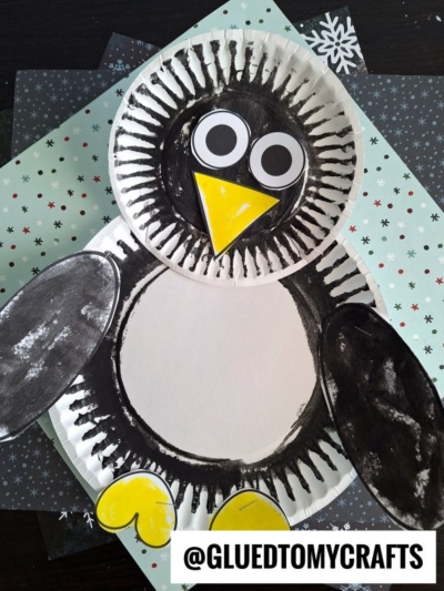 A paper plate penguin craft colored in black, white, and yellow features large googly eyes and sits on layered snowflake-patterned papers. @GLUEDTOMYCRAFTS is written at the bottom.