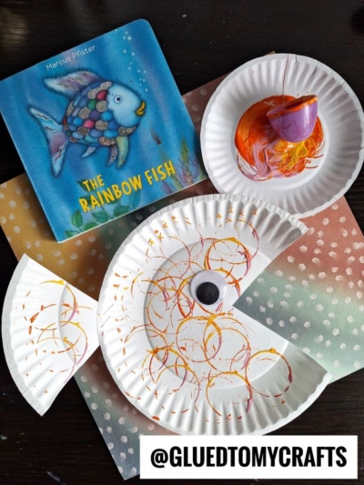 A childrens book titled The Rainbow Fish sits beside a fish craft made from a painted, cut paper plate and a googly eye, inspired by Plastic Egg Stamp Crafts, all on a polka dot background. @GLUEDTOMYCRAFTS is written at the bottom.