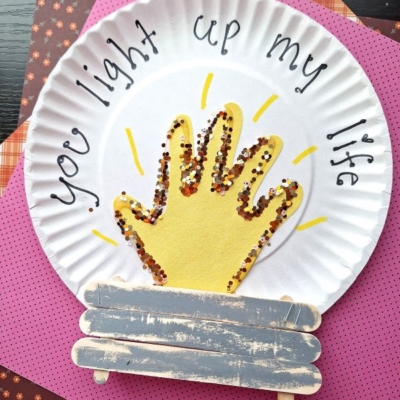 A paper plate craft features a yellow painted handprint with glitter, resembling a campfire, on a paper plate. “You light up my life” is written around the top, and painted craft stick logs are at the bottom.