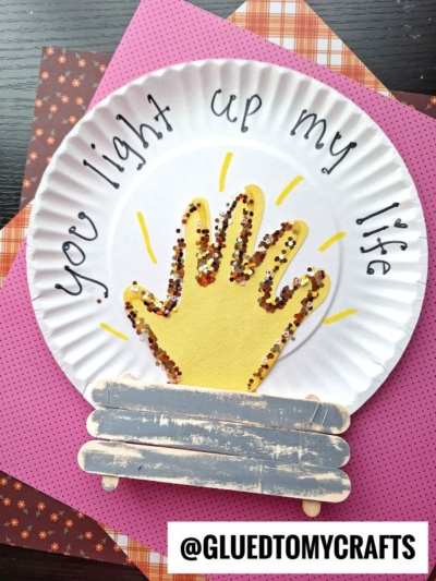 A paper plate craft features a yellow painted handprint with glitter, resembling a campfire, on a paper plate. “You light up my life” is written around the top, and painted craft stick logs are at the bottom.
