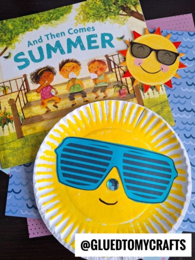 A childrens book titled And Then Comes Summer is displayed with two paper plate sun crafts: one with blue sunglasses and a smile, and another smaller sun with a pink rim and black sunglasses. @gluedtomycrafts appears at the bottom.