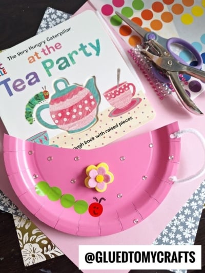 A pink paper plate purse craft with a flower, ladybug, and hints of a HUNGRY CATERPILLAR CRAFT is shown on a table with a book, colorful dot stickers, purple beads, and crafting supplies. @GLUEDTOMYCRAFTS is at the bottom.