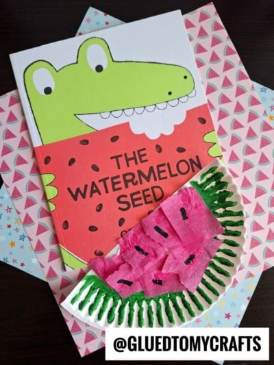 A children’s book titled The Watermelon Seed with a cartoon crocodile on the cover sits on colorful paper, next to a handmade watermelon craft—perfect for summer craft ideas using green yarn and pink tissue paper.