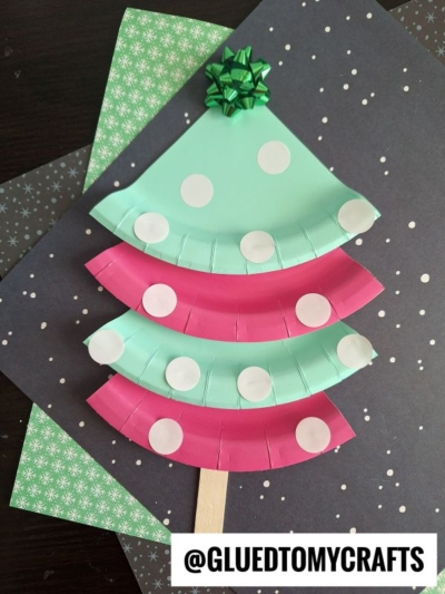 A paper plate dot sticker Christmas tree craft, painted in teal and pink, features white dot stickers, a green bow topper, and a wood stick trunk, all displayed on patterned paper.