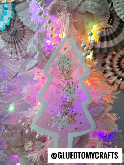 A pink Christmas tree ornament with silver snowflakes and gold glitter hangs on a pastel pink stained glass Christmas tree adorned with flowers, ornaments, and soft white lights. @GLUEDTOMYCRAFTS is displayed at the bottom.