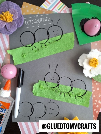 Two black paper circles on gray paper, partly covered with green tissue, resemble caterpillars with smiling faces and antennae. Plastic Egg Stamp Crafts and other supplies are arranged around the colorful artwork.